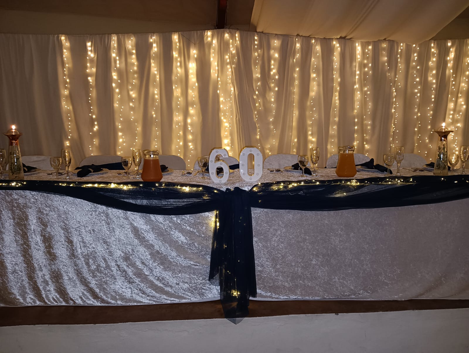 Elegant 60th birthday celebration main table with white draping, fairy lights, black table runner and illuminated number 60 at Mayerskraal Wedding Venue