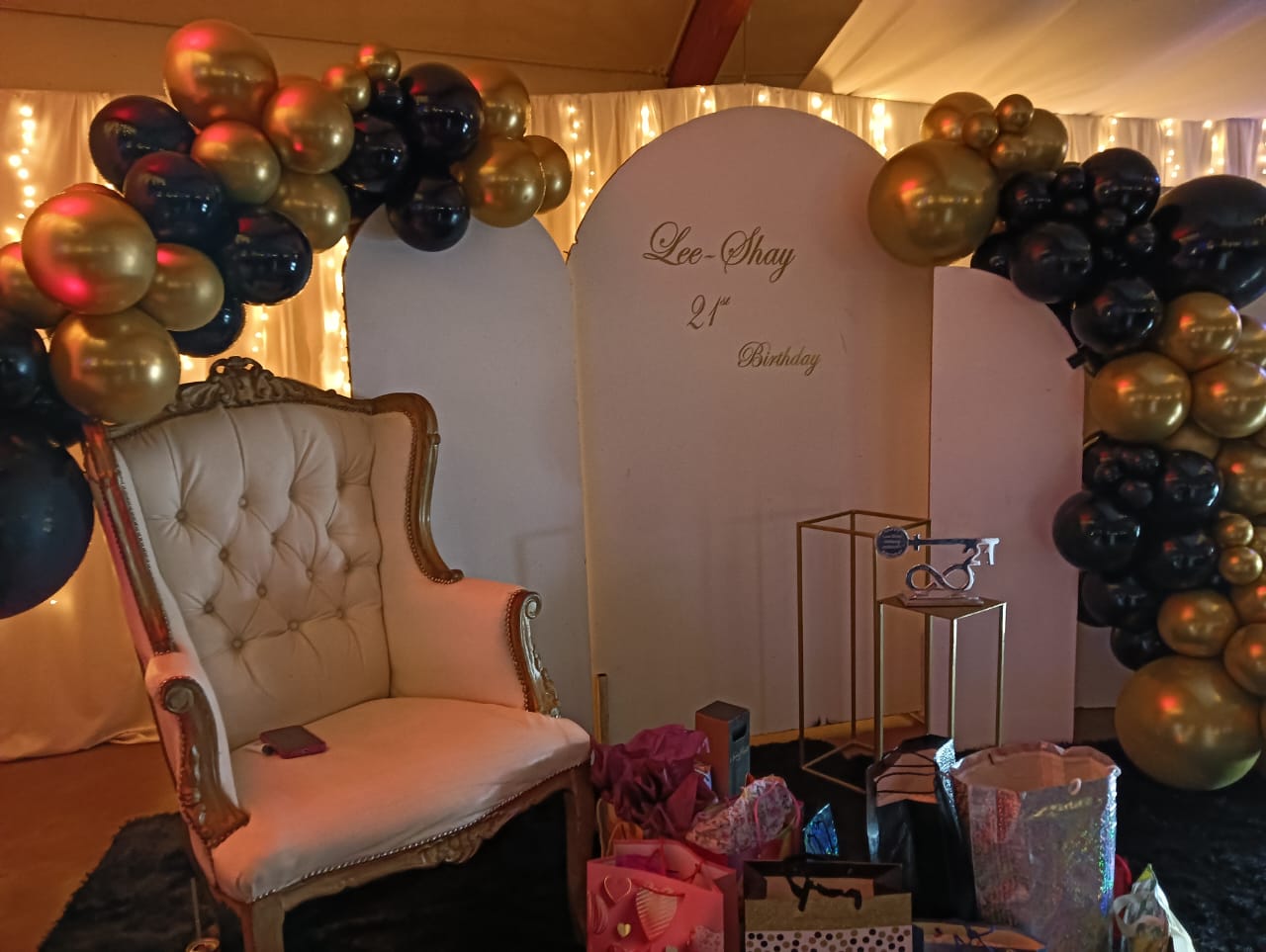 Elegant birthday celebration setup at Mayerskraal featuring a statement chair, illuminated backdrop, gold and black balloon arrangement, personalised signage and gift table.