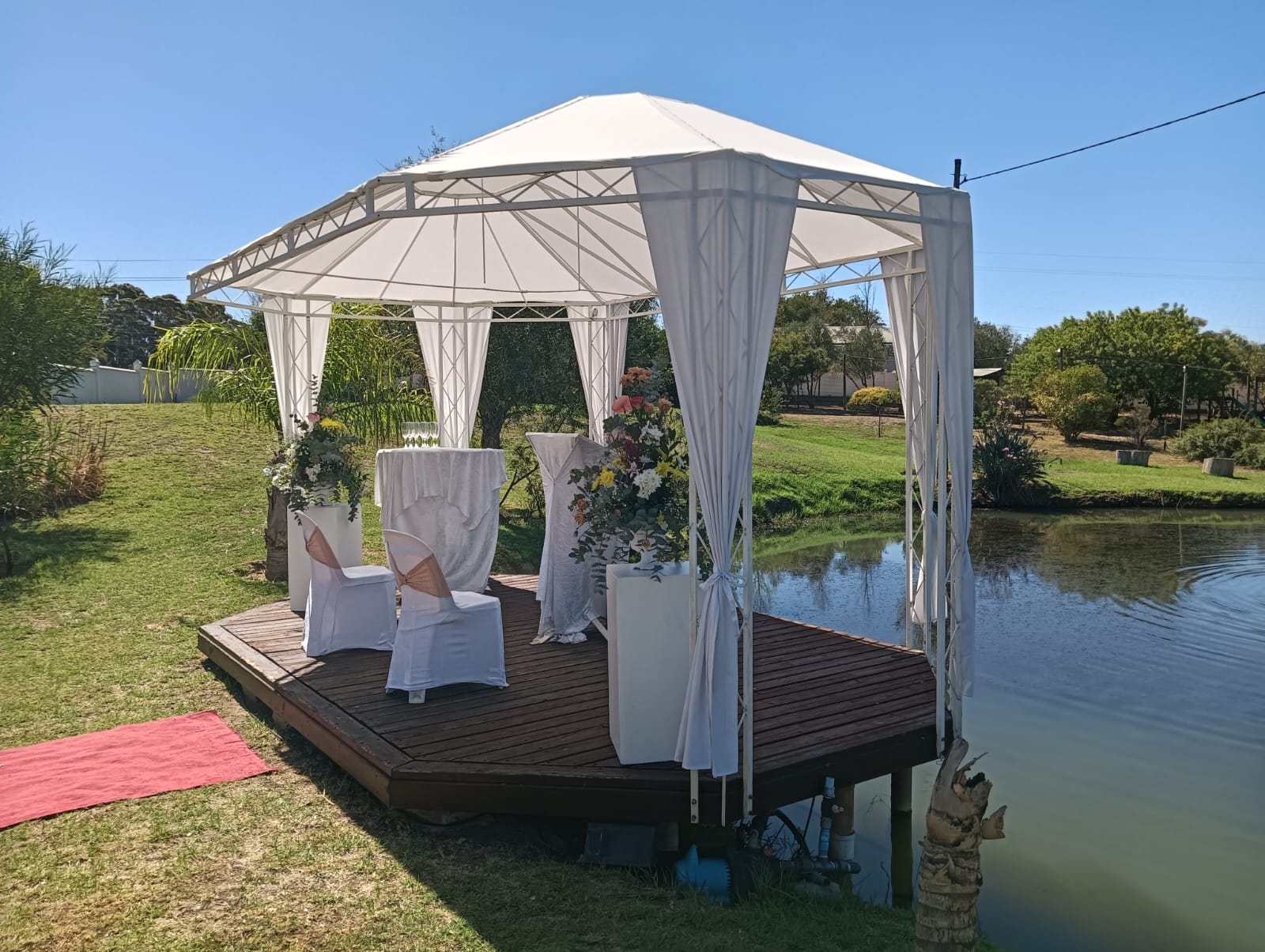 Elegant outdoor wedding ceremony setup on a wooden deck beside the pond at Mayerskraal, with white draping, floral arrangements and mountain views.
