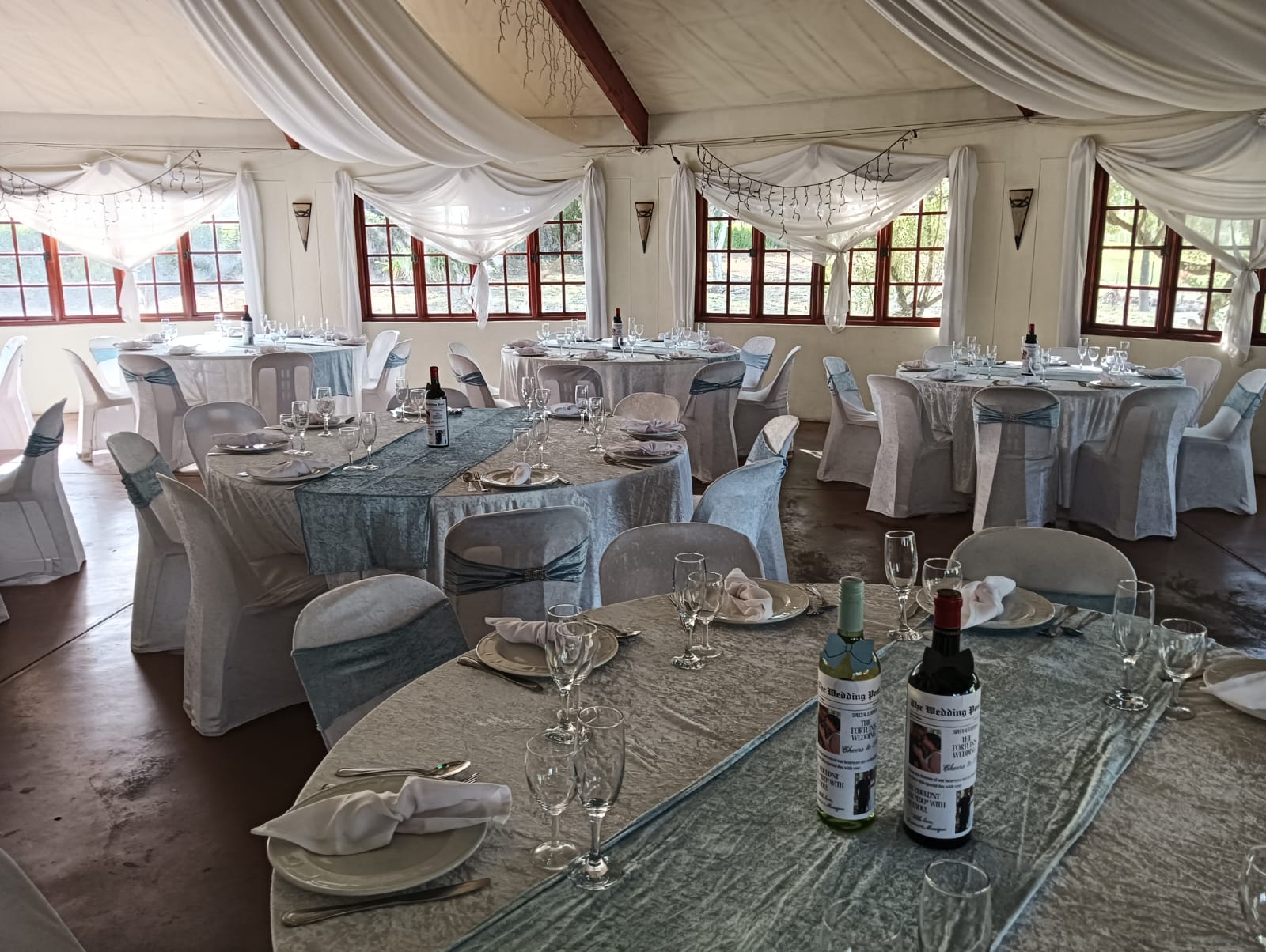 Beautifully dressed wedding reception tables at Mayerskraal, featuring silver-blue tablecloths, matching chair sashes, floral-style draping and personalised wine bottles.