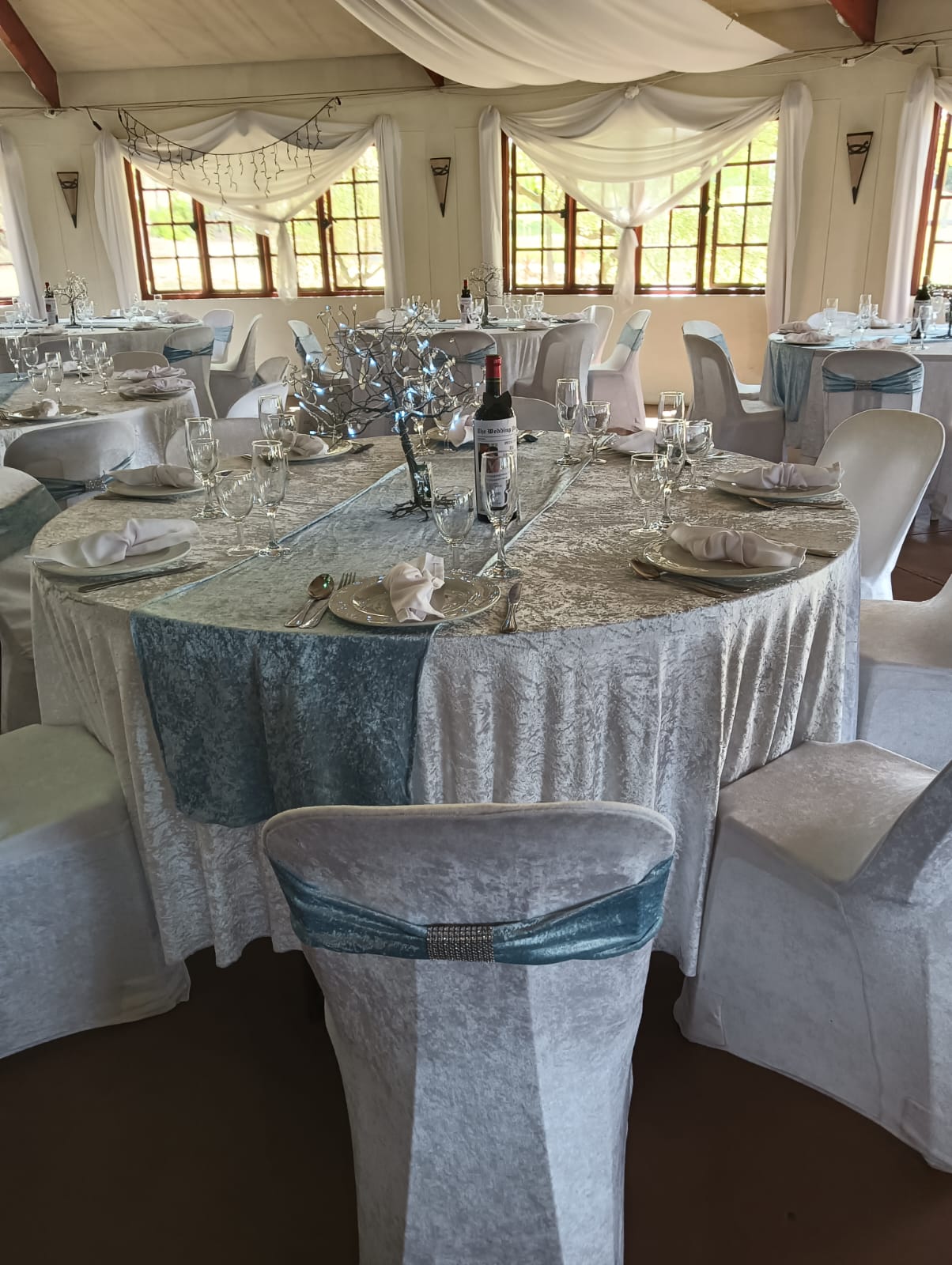 Elegant wedding reception table at Mayerskraal with silver-blue tablecloths, white napkins, glassware, personalised wine bottles and illuminated branch centerpieces.