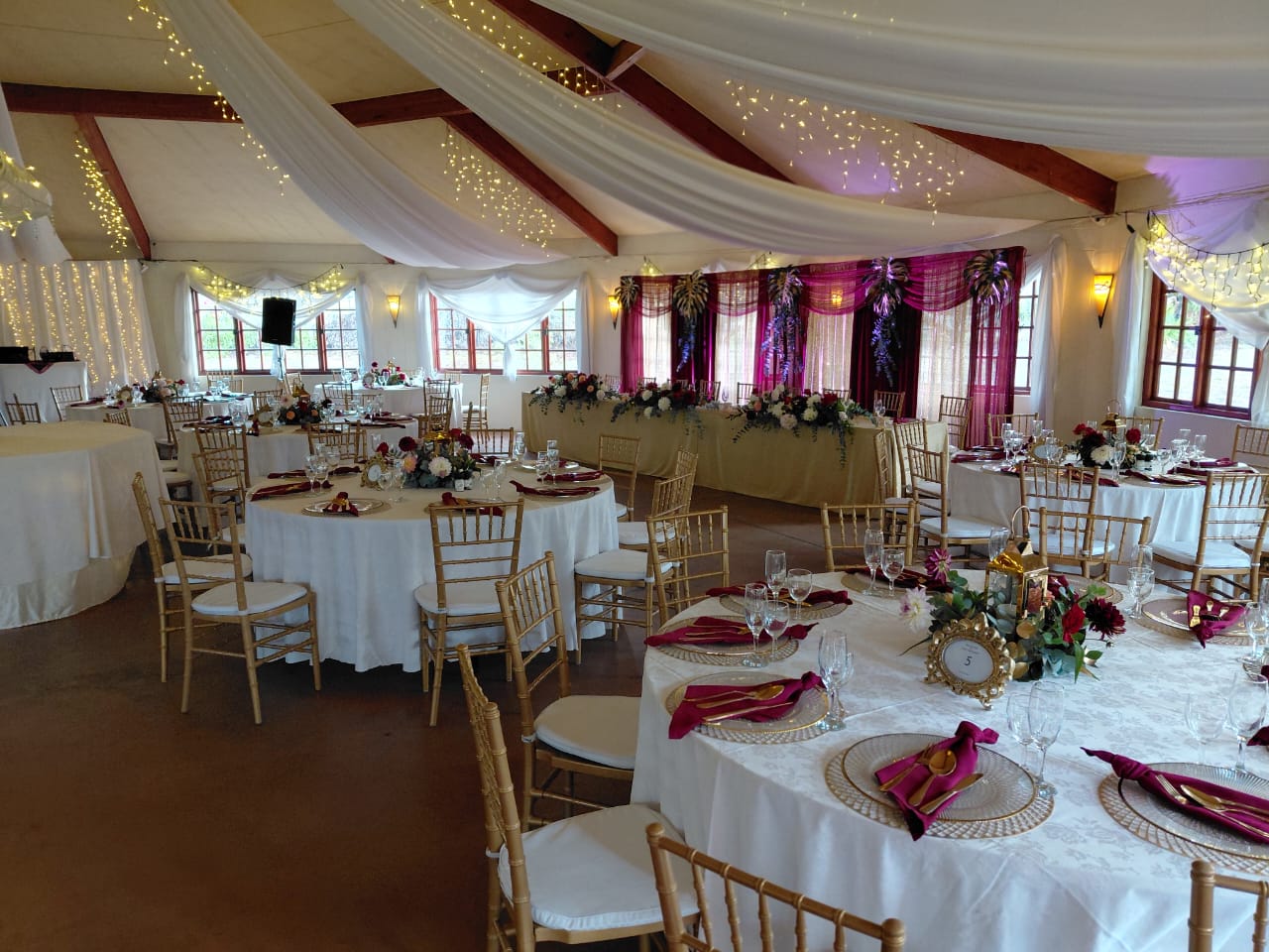 Elegant wedding reception with round tables, gold Chiavari chairs, floral centerpieces, burgundy napkins and draped ceiling décor at Mayerskraal Wedding Venue