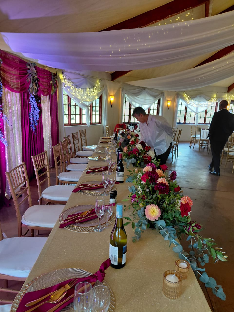 Long wedding reception table with colourful floral centerpiece, gold place settings, burgundy napkins and candles at Mayerskraal Wedding Venue
