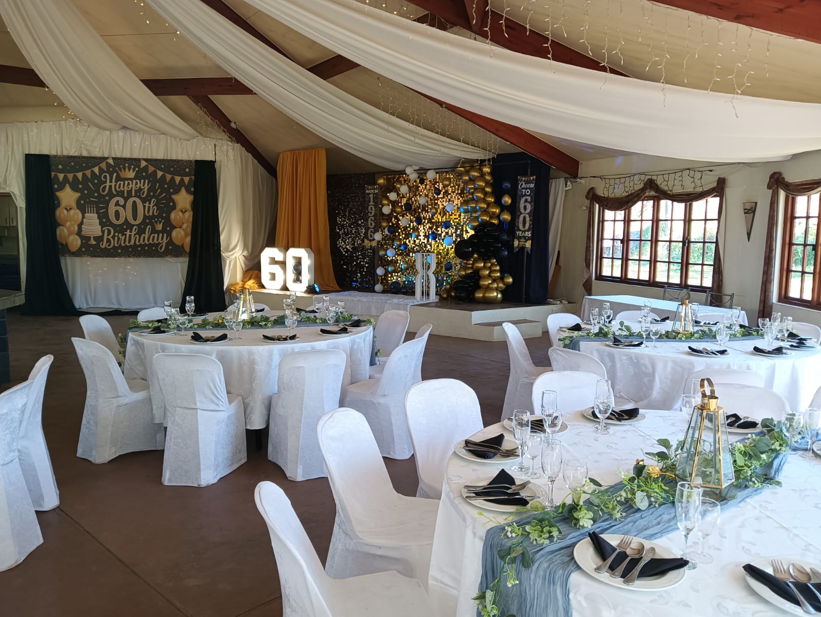 60th Birthday celebration with white tables, gold and black décor, balloon backdrop and draped ceiling at Mayerskraal Events Venue